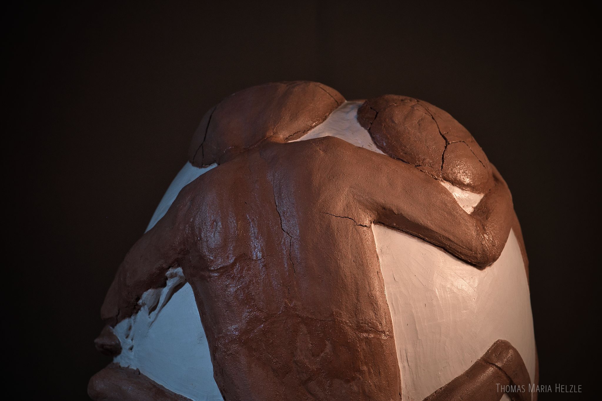 Closeup of the Lovers sculpture, showing the upper back of the heavier figure with cracks and overgrown areas. The second figure is only partially visible. The basis is an egg-shape made of plaster with the two figures modelled from dark brown-red clay in relief on top. The background is dark grey.